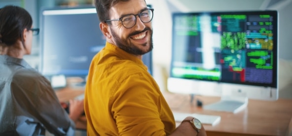Ein Mann dreht sich von seinem Schreibtisch zu der Kamera um und lächelt herzlich. Er arbeitet gerade mit seinem Computer - darauf zu sehen ist eine Software zur Kundenverwaltung.