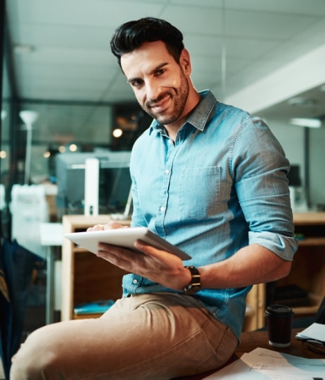Ein junger Unternehmer lehnt an einem Schreibtisch in seinem Büroräumen. Er hält ein Tablet in der Hand und lächelt zufrieden, weil seine IT-Ausstattung ihm sehr viel Arbeit abnimmt.