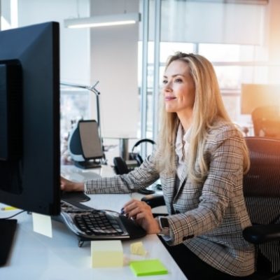 Eine junge Frau sitzt an Ihrem Schreibtisch - dieser ist mit meherern Bildschirmen, Maus und Tastatur ausgestattet.