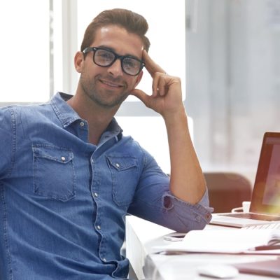 Ein junger Unternehmer sitzt an seinem Schreibtisch und lächelt entspannt in die Kamera. Er ist zufrieden mit seinem IT-Dienstleister, weil er durch diesen mehr Zeit für sein eigenes Unternehmen hat.